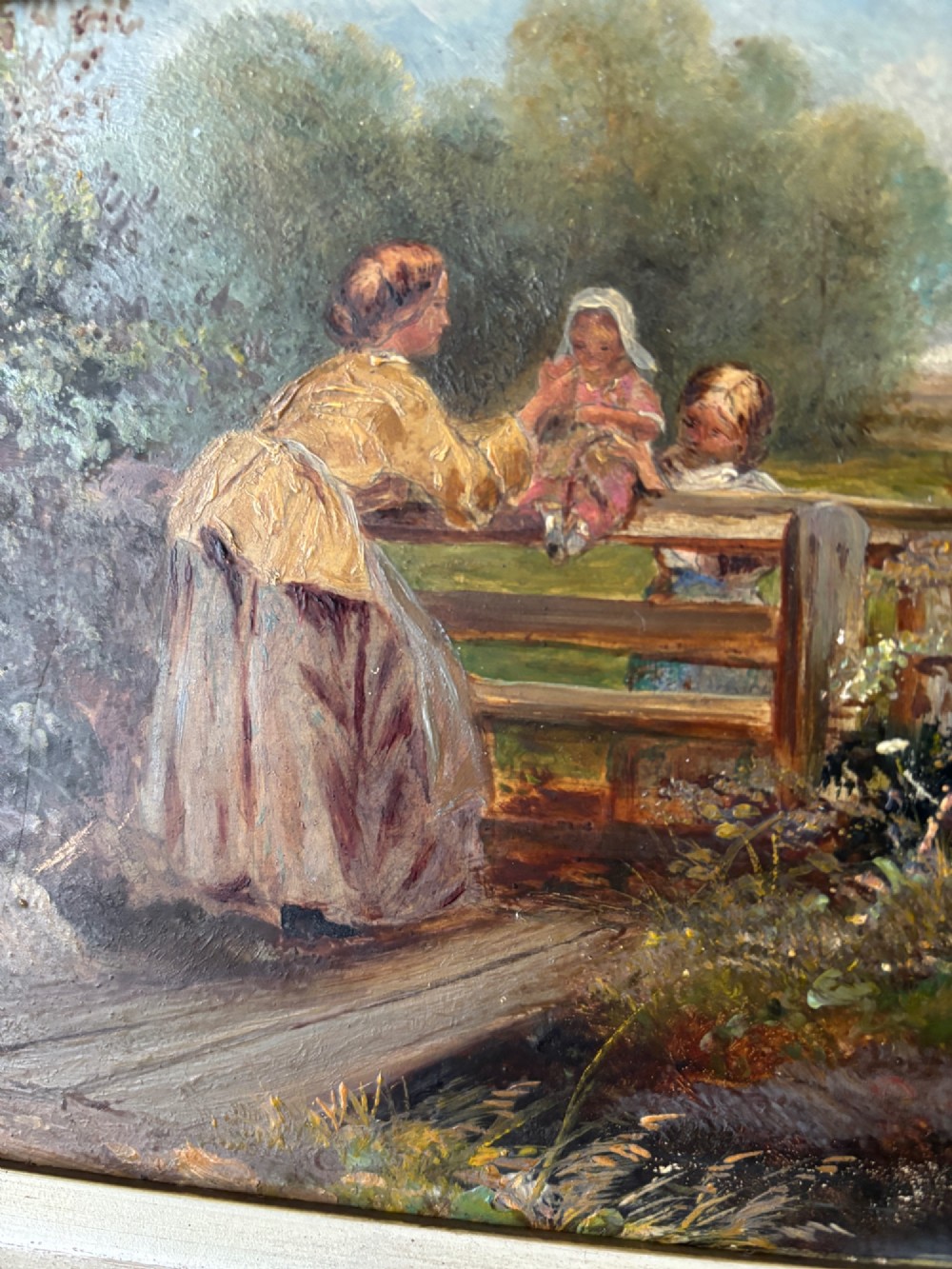 A small 19th-century style oil painting on board depicting a woman and children near a rustic wooden fence, set in a wide gilded frame. View III