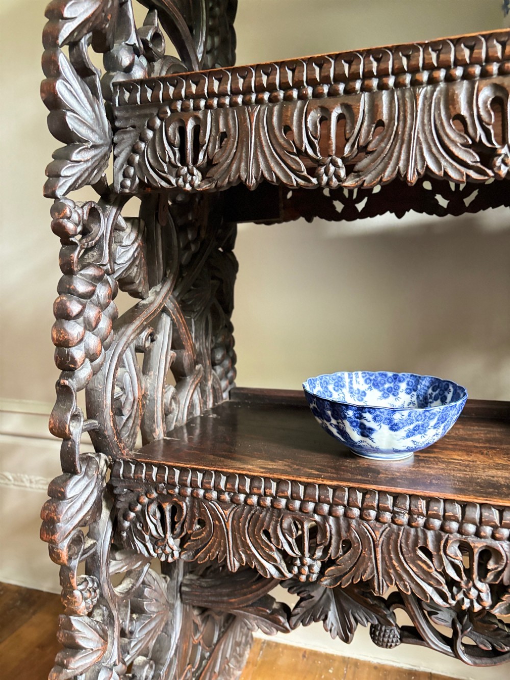 A tall mid-19th-century Anglo-Indian padauk wood shelving unit with three open tiers featuring deeply carved foliate aprons and intricate side supports. View IV