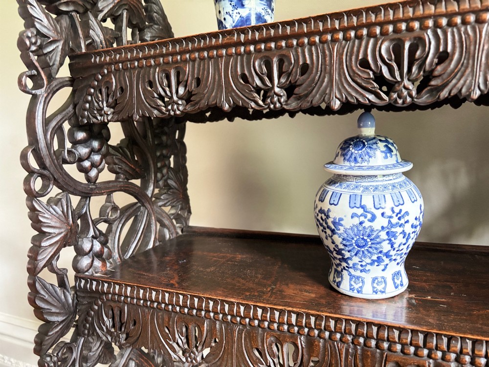 A tall mid-19th-century Anglo-Indian padauk wood shelving unit with three open tiers featuring deeply carved foliate aprons and intricate side supports. View V