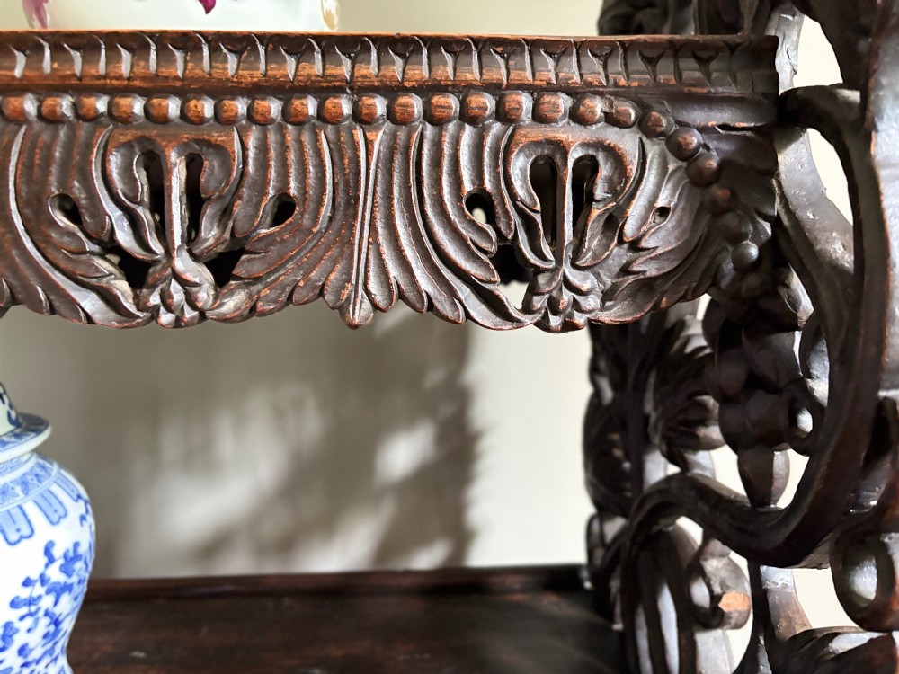 A tall mid-19th-century Anglo-Indian padauk wood shelving unit with three open tiers featuring deeply carved foliate aprons and intricate side supports. View VII