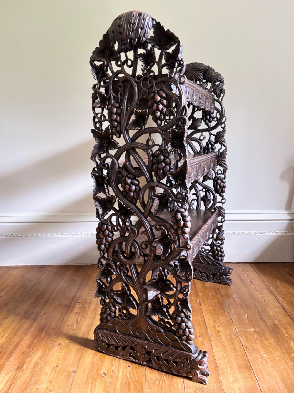 A tall mid-19th-century Anglo-Indian padauk wood shelving unit with three open tiers featuring deeply carved foliate aprons and intricate side supports. View IX