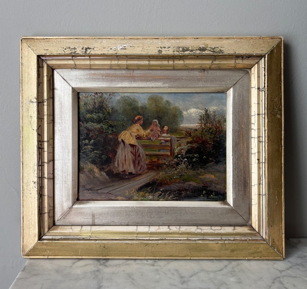 A small 19th-century style oil painting on board depicting a woman and children near a rustic wooden fence, set in a wide gilded frame.
