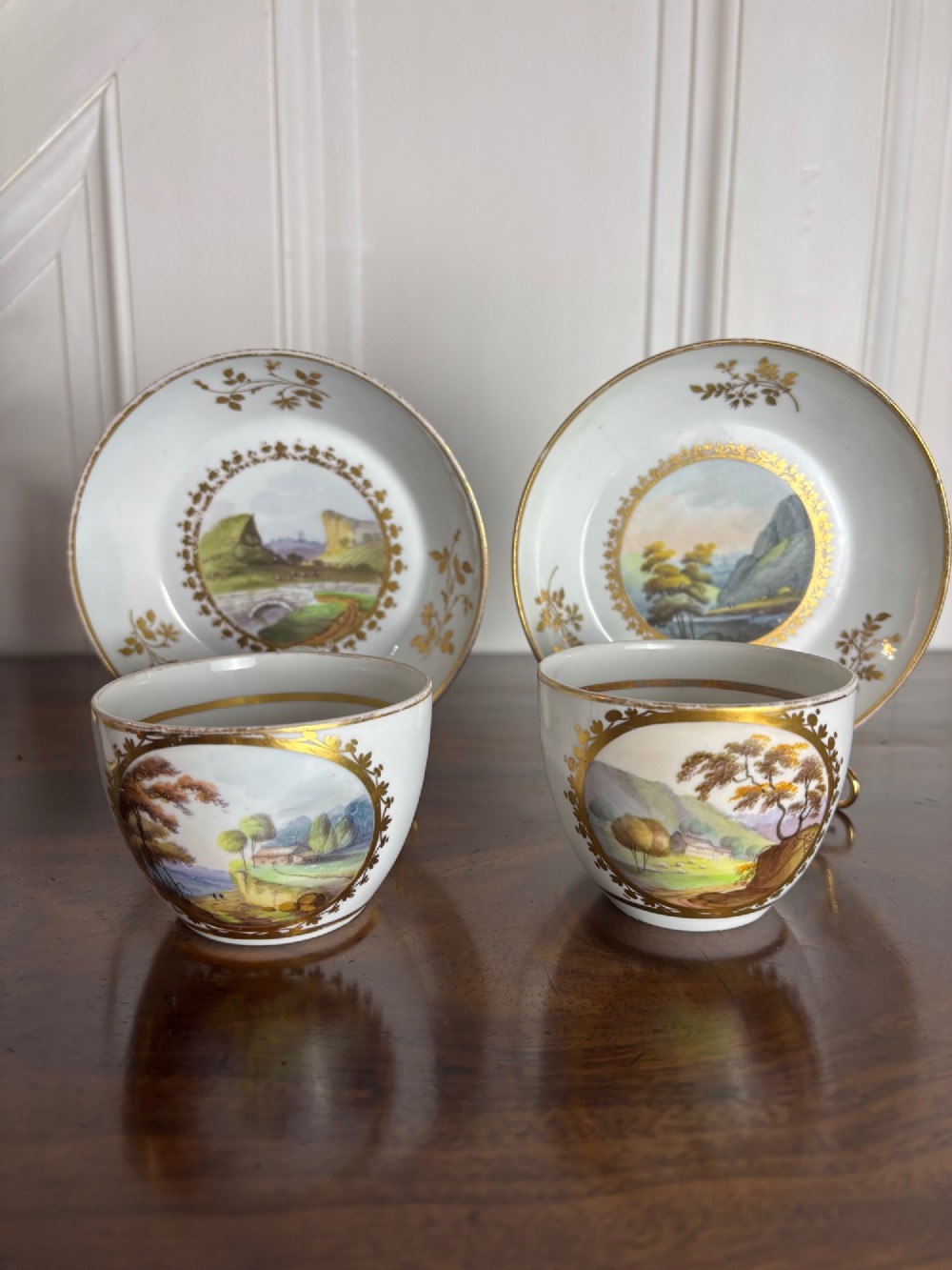 A pair of white Georgian porcelain teacups and saucers with hand-painted landscape scenes enclosed within gilded foliate borders.