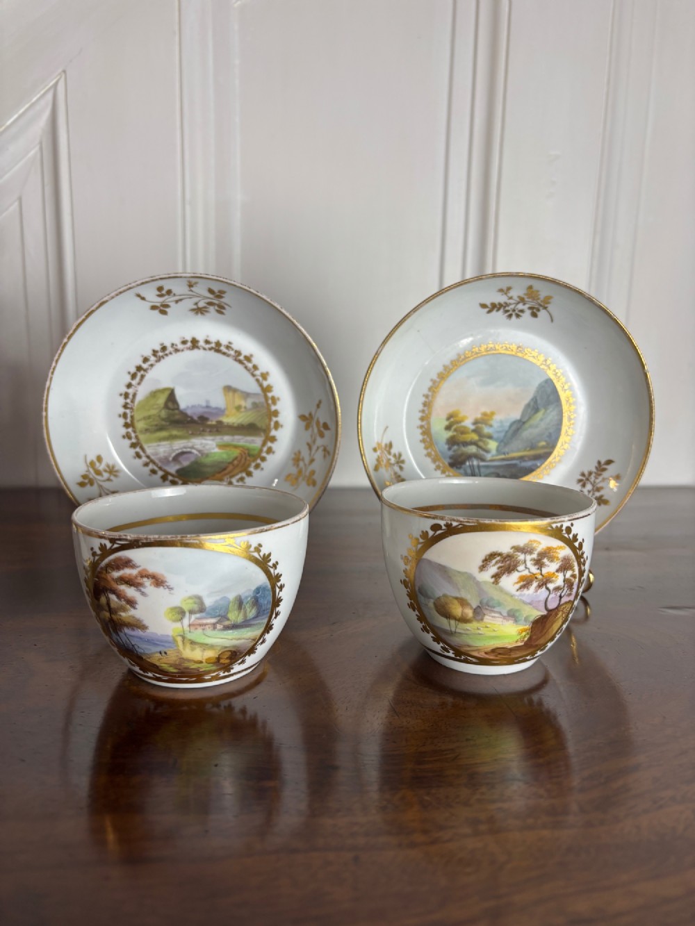 A pair of white Georgian porcelain teacups and saucers with hand-painted landscape scenes enclosed within gilded foliate borders. View II