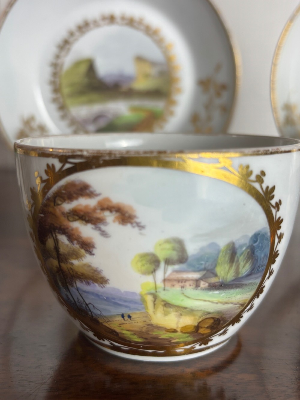 A pair of white Georgian porcelain teacups and saucers with hand-painted landscape scenes enclosed within gilded foliate borders. View III