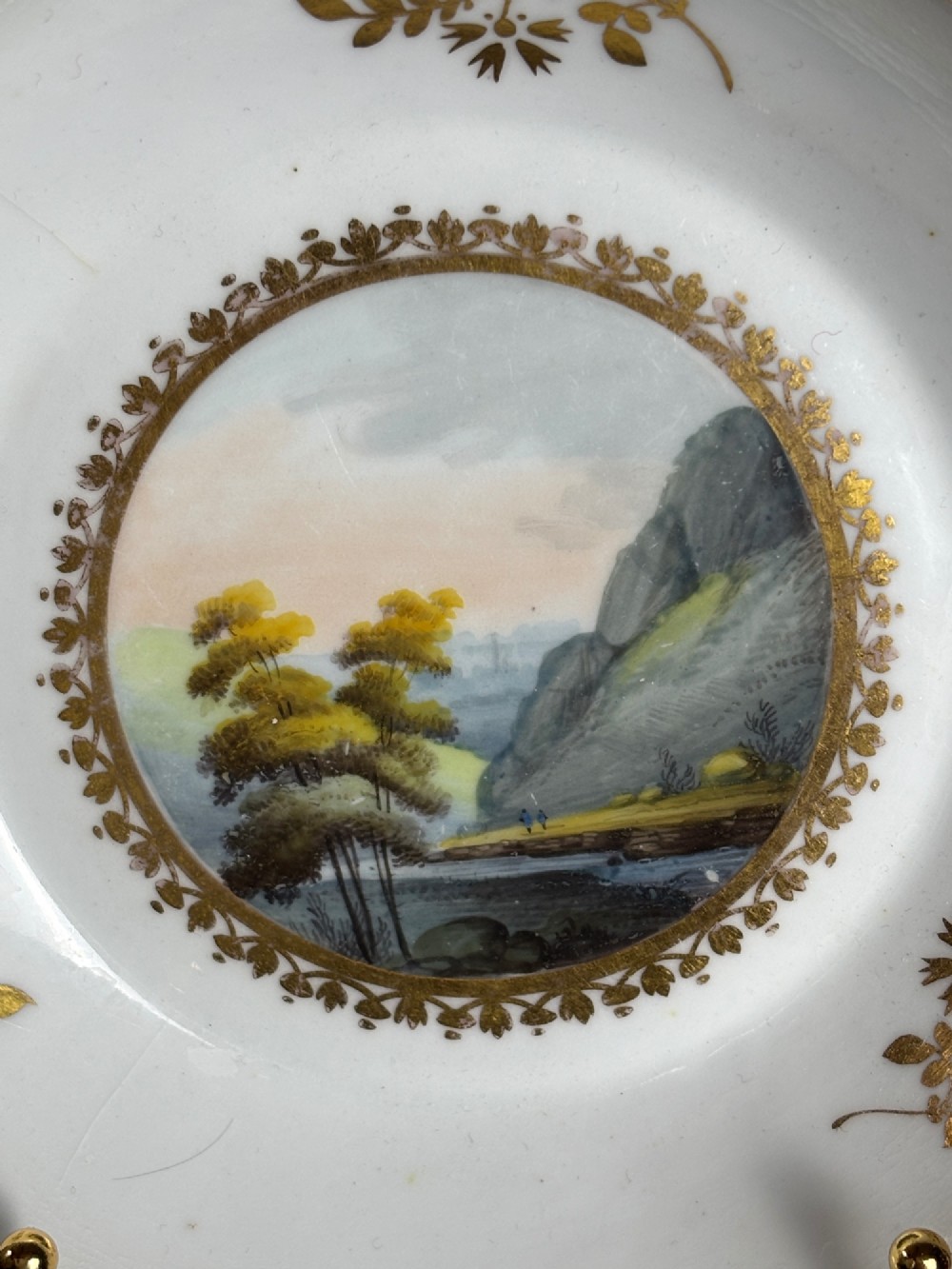 A pair of white Georgian porcelain teacups and saucers with hand-painted landscape scenes enclosed within gilded foliate borders. View V