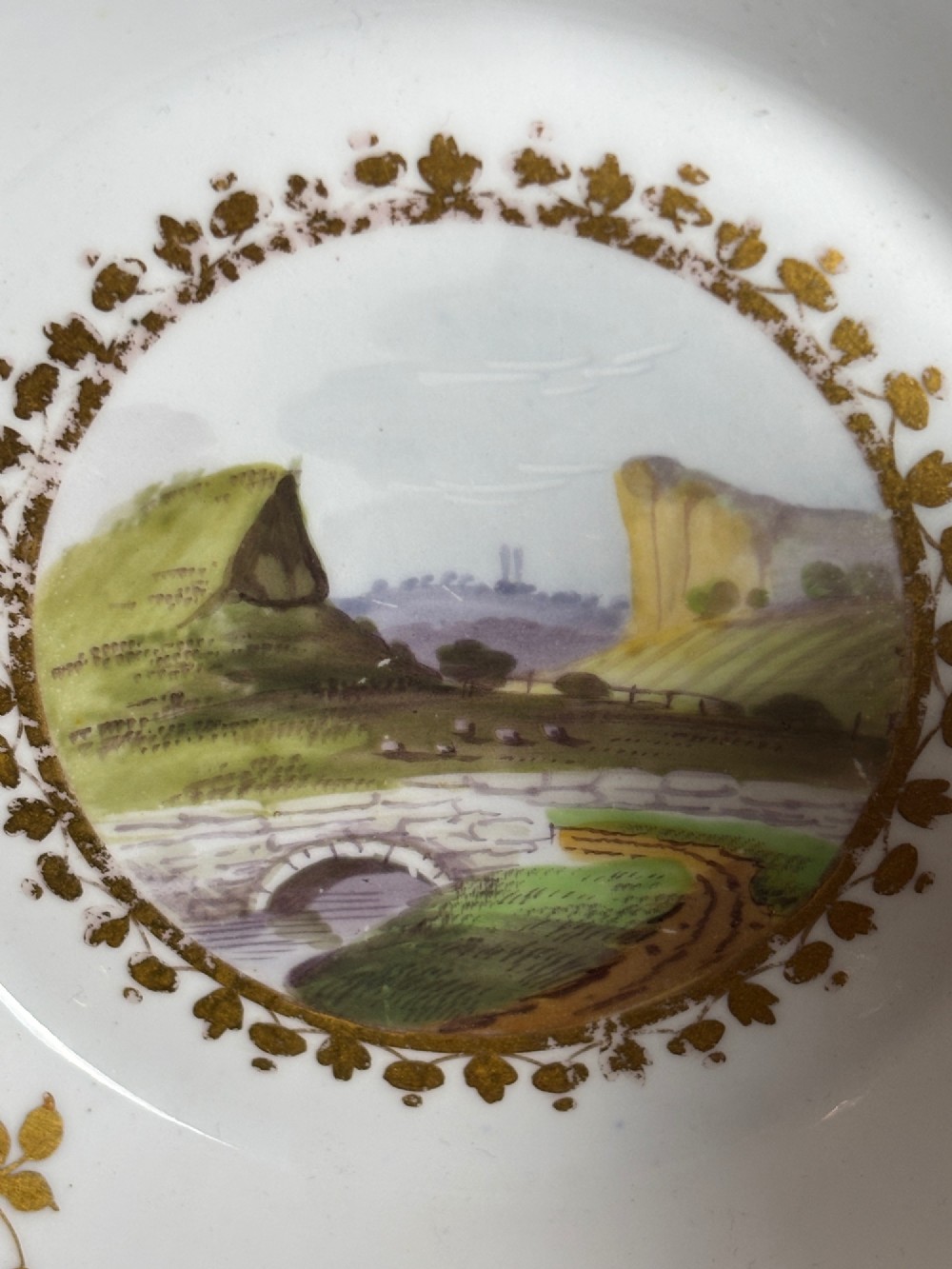 A pair of white Georgian porcelain teacups and saucers with hand-painted landscape scenes enclosed within gilded foliate borders. View VI