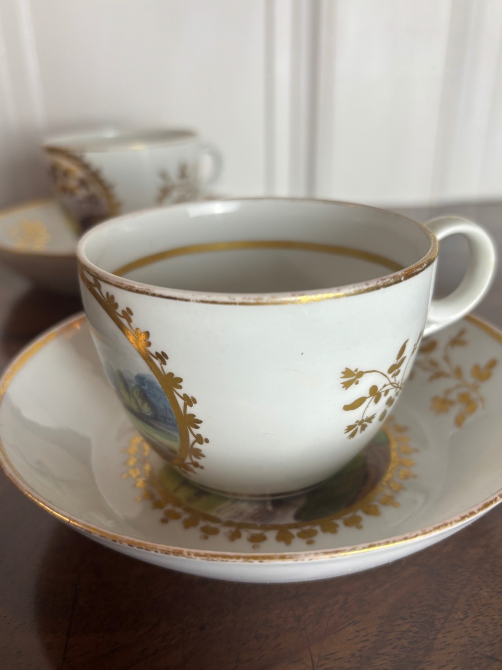 A pair of white Georgian porcelain teacups and saucers with hand-painted landscape scenes enclosed within gilded foliate borders. View VIII
