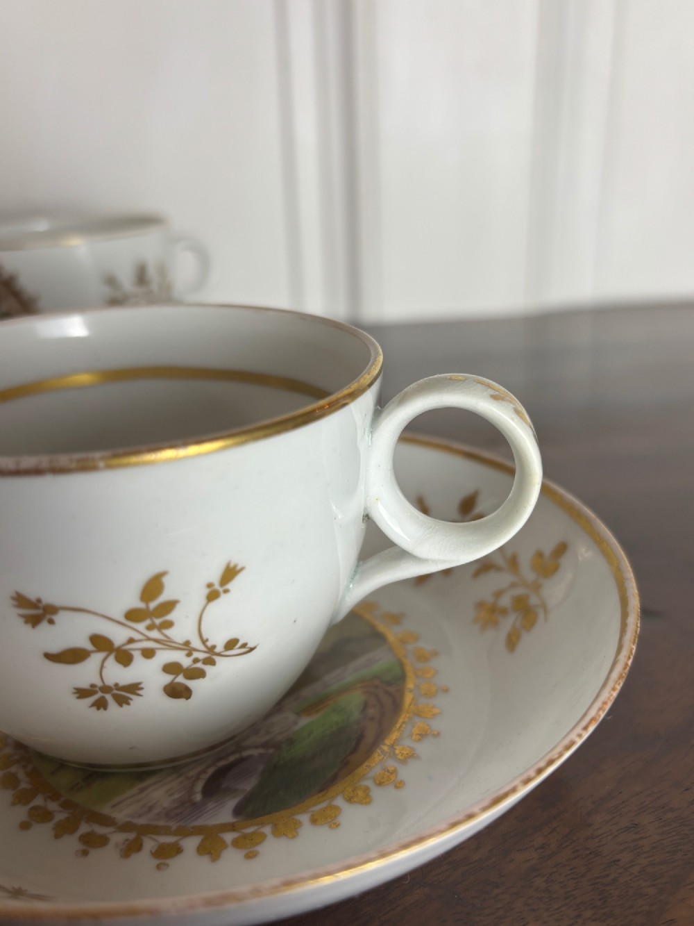 A pair of white Georgian porcelain teacups and saucers with hand-painted landscape scenes enclosed within gilded foliate borders. View IX