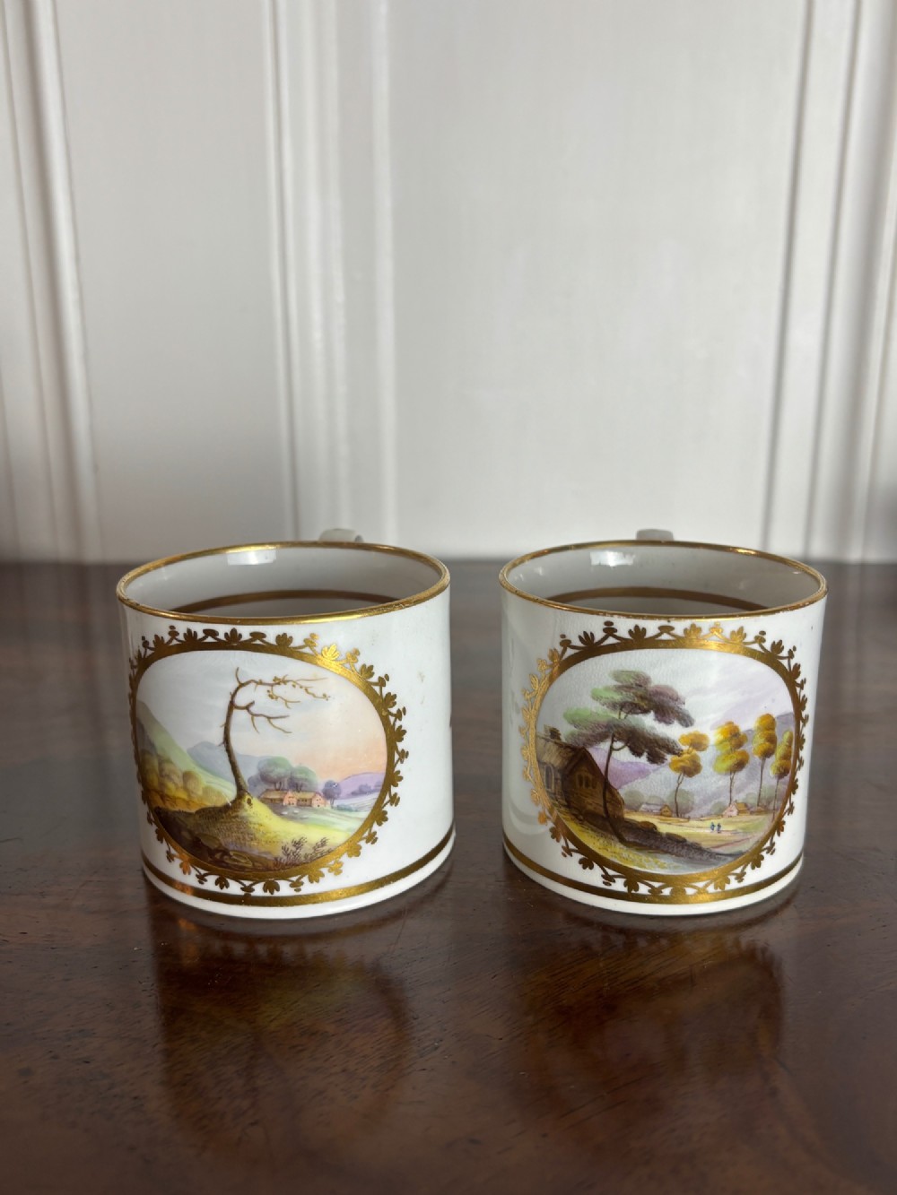 A pair of early nineteenth-century white porcelain coffee cans featuring hand-painted oval landscape vignettes framed with gilded borders.