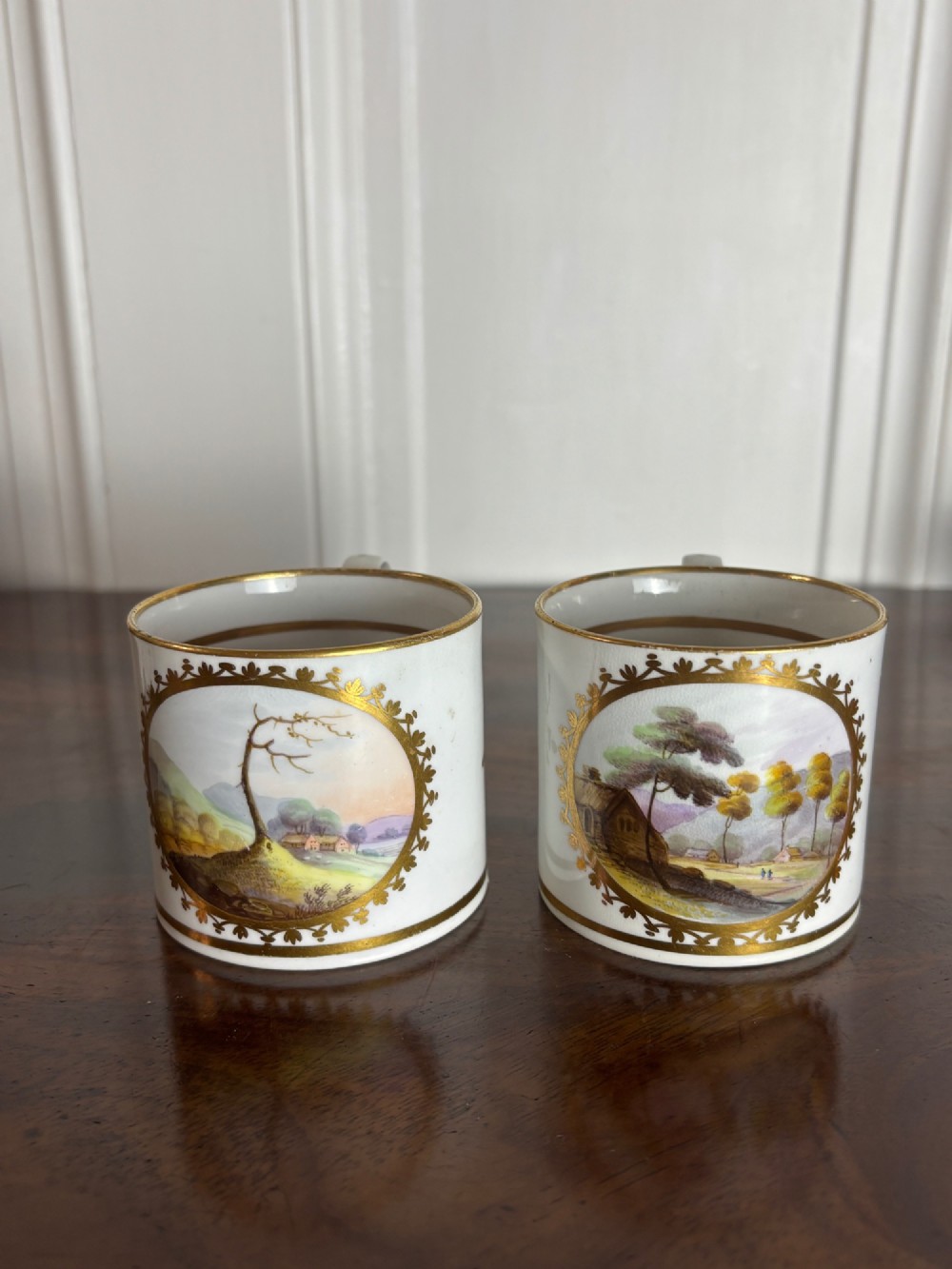 A pair of early nineteenth-century white porcelain coffee cans featuring hand-painted oval landscape vignettes framed with gilded borders. View II