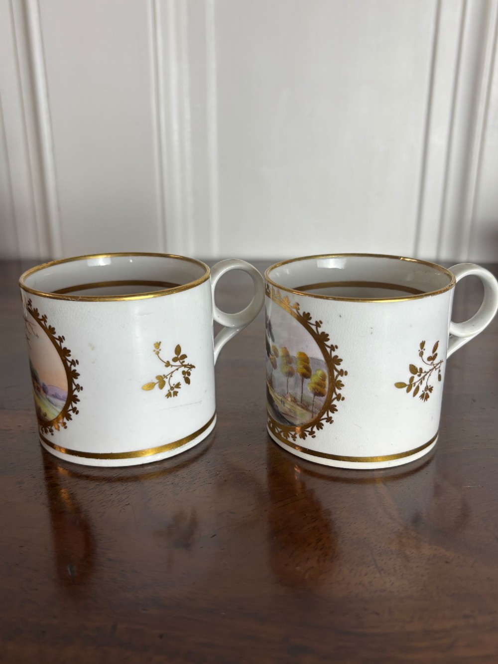 A pair of early nineteenth-century white porcelain coffee cans featuring hand-painted oval landscape vignettes framed with gilded borders. View III