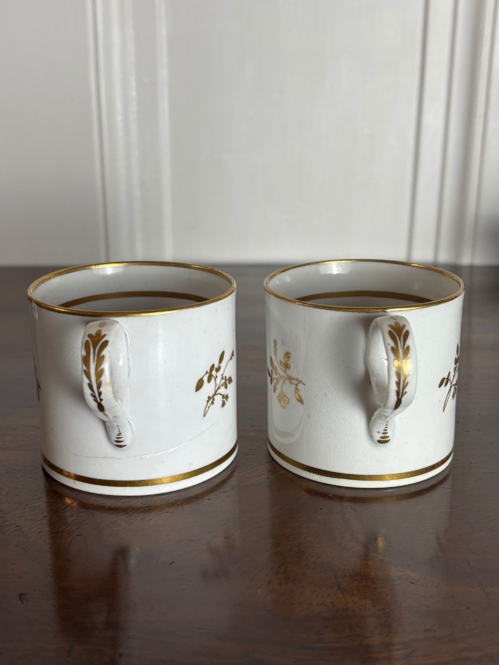 A pair of early nineteenth-century white porcelain coffee cans featuring hand-painted oval landscape vignettes framed with gilded borders. View V