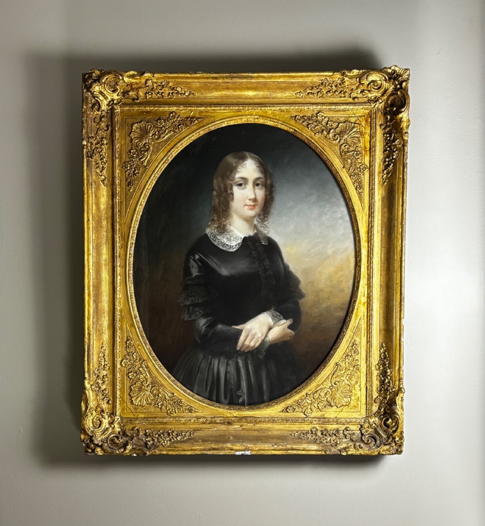 An oil portrait of a dark-haired woman in a black dress housed within an oval spandrel and a heavily carved gilt frame.