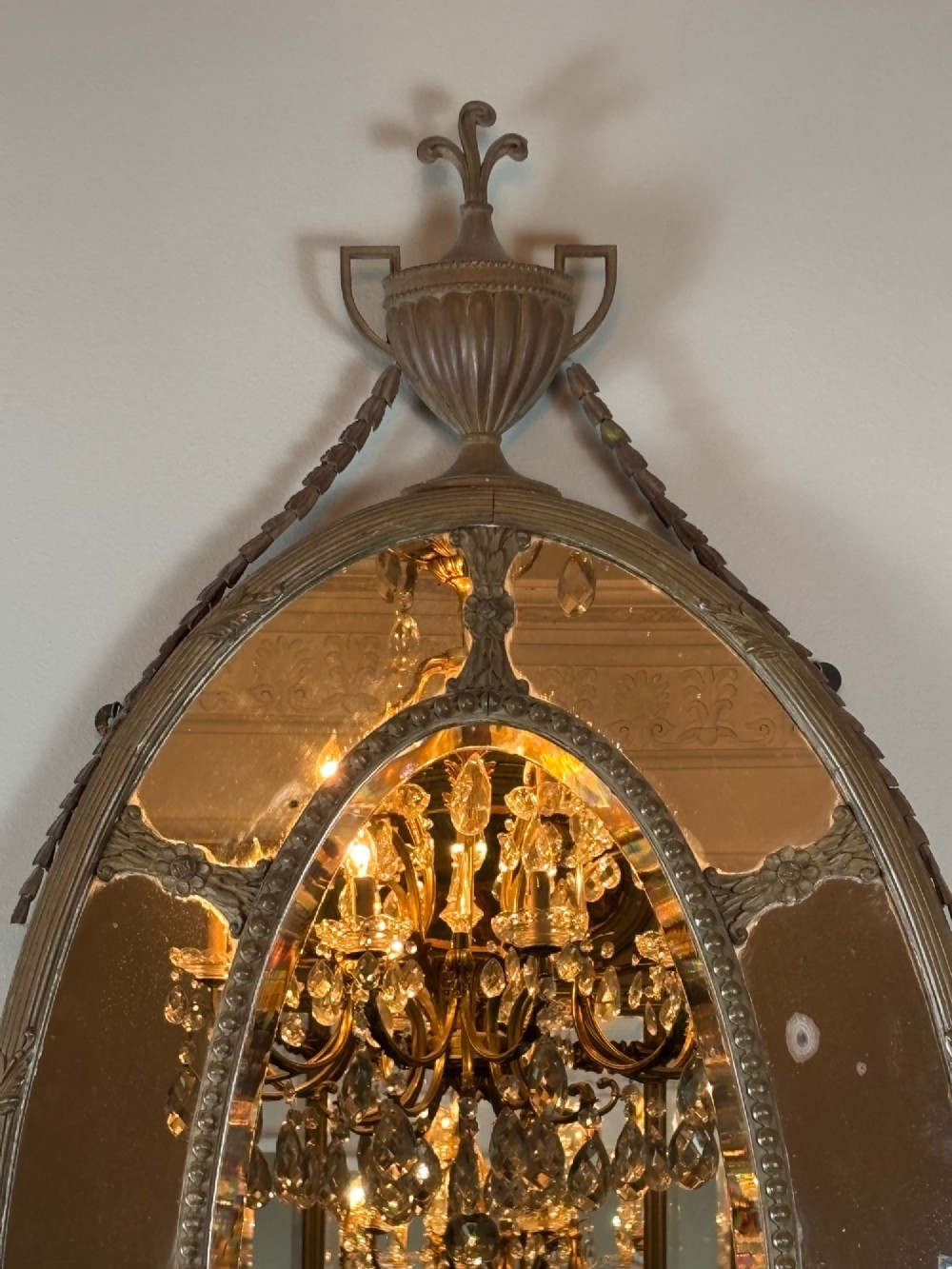 An oval late Victorian wall mirror featuring a central mirror plate surrounded by border glass, topped with a classical urn and draped swag cresting. View II