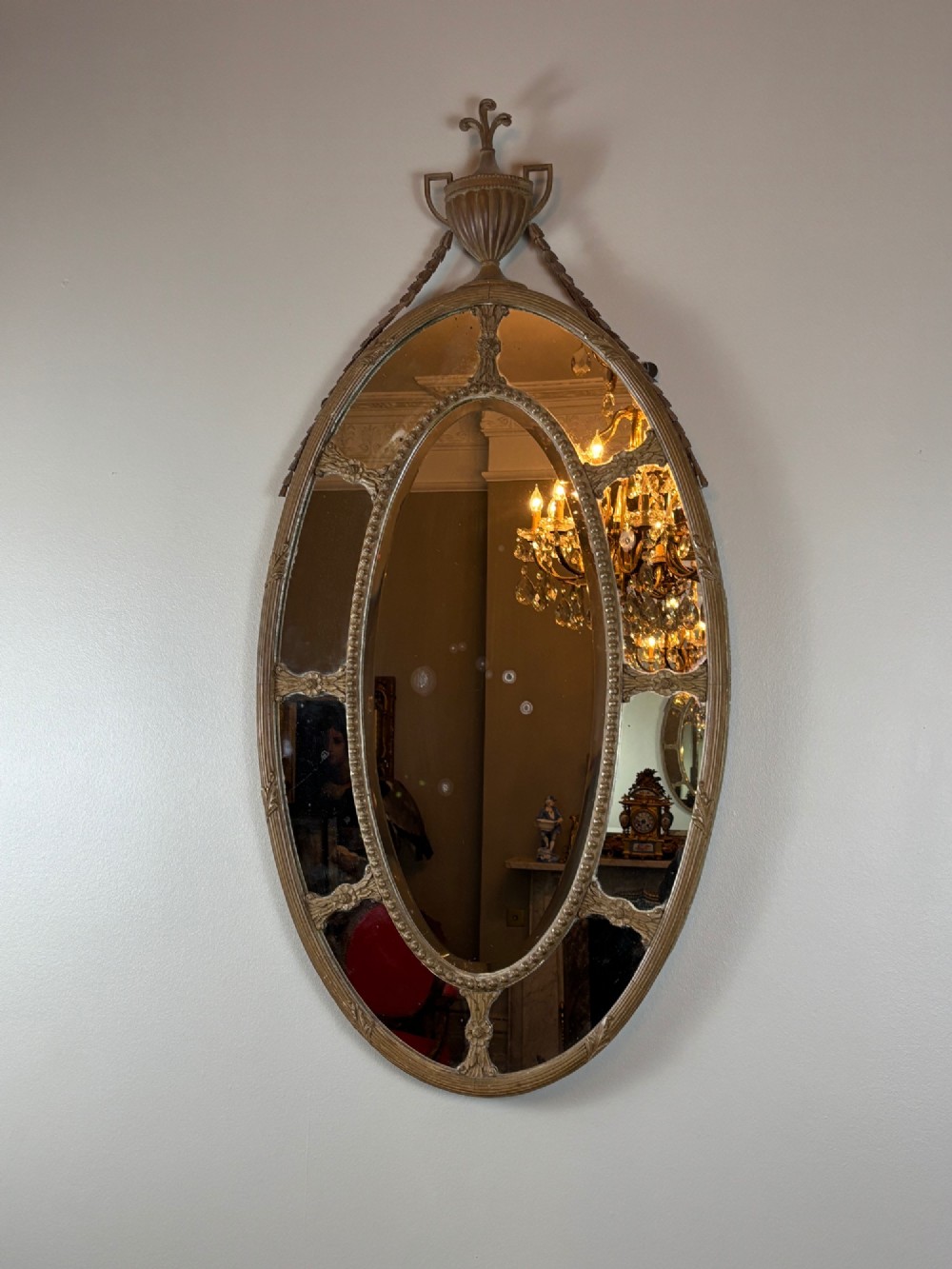 An oval late Victorian wall mirror featuring a central mirror plate surrounded by border glass, topped with a classical urn and draped swag cresting. View III