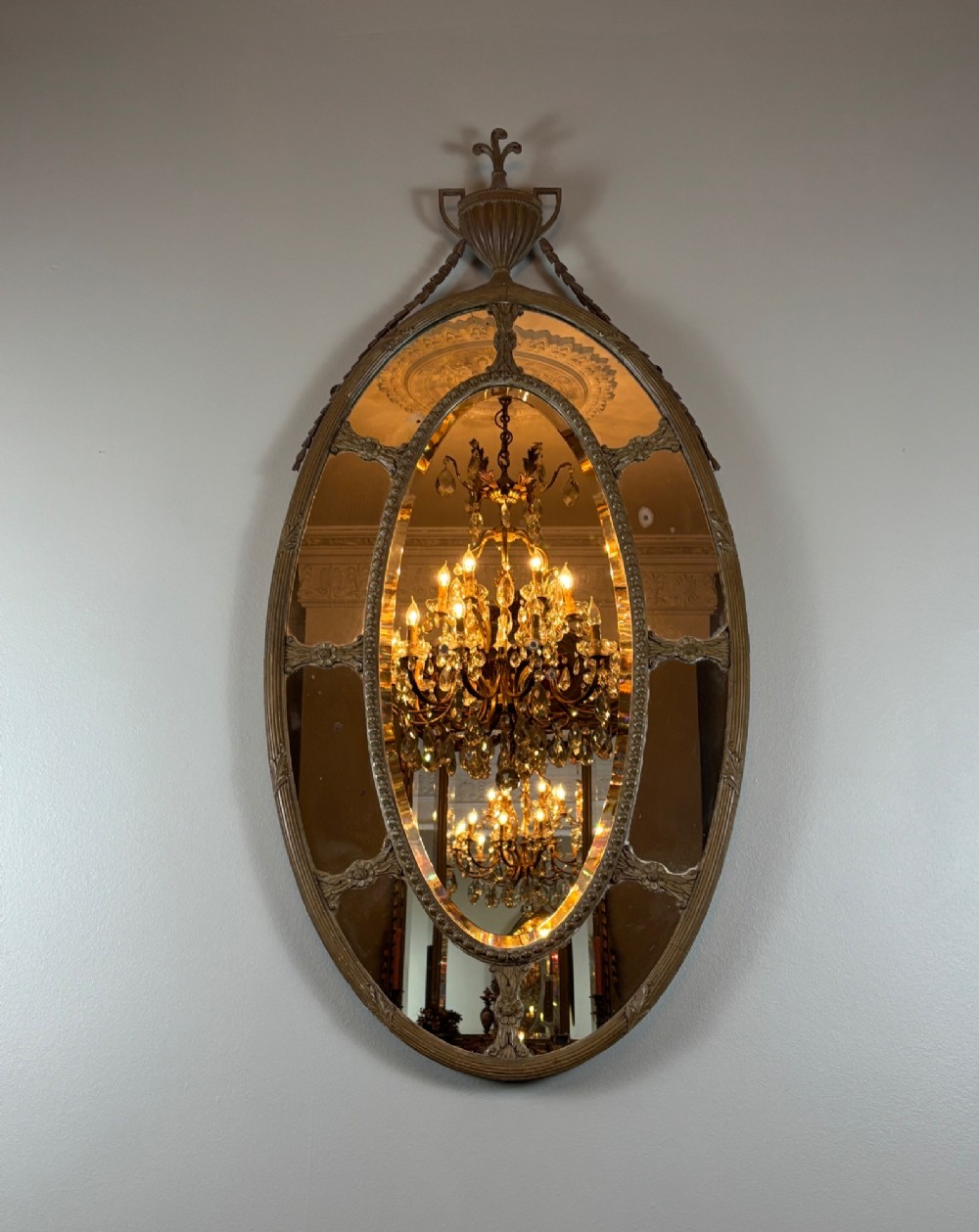 An oval late Victorian wall mirror featuring a central mirror plate surrounded by border glass, topped with a classical urn and draped swag cresting.