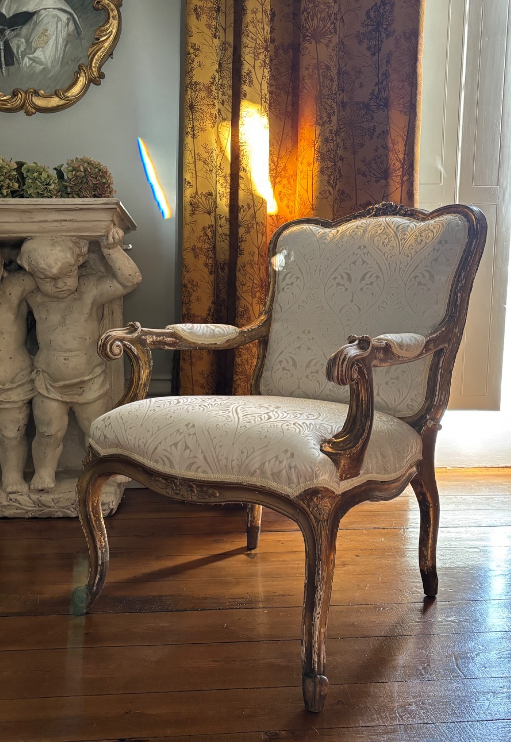 A Louis XV style open armchair featuring a distressed giltwood frame with cabriole legs, padded manchettes, and cream patterned silk damask upholstery.