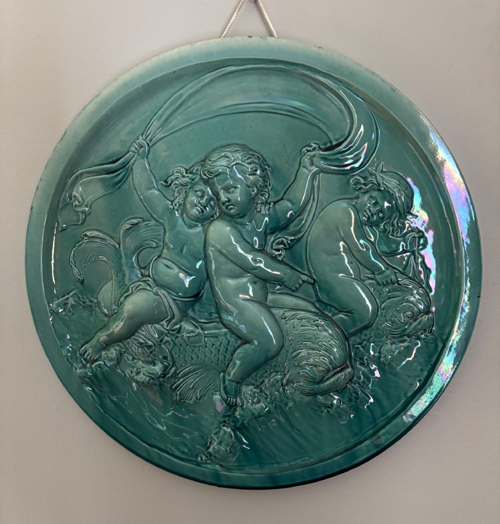 A circular turquoise-glazed ceramic plaque depicting cherubs sculpted in high relief holding flowing drapery.