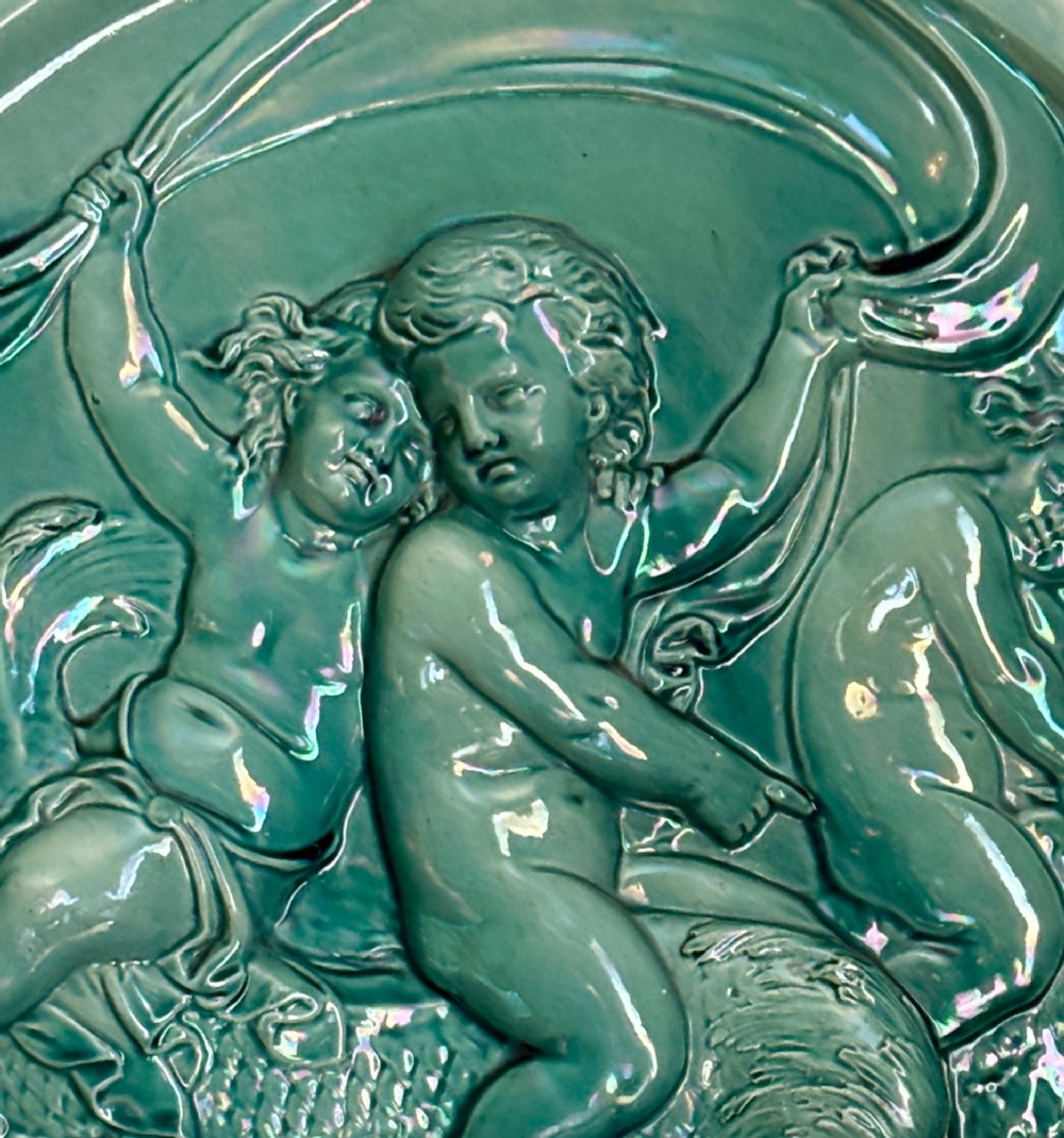 A circular turquoise-glazed ceramic plaque depicting cherubs sculpted in high relief holding flowing drapery. View IV