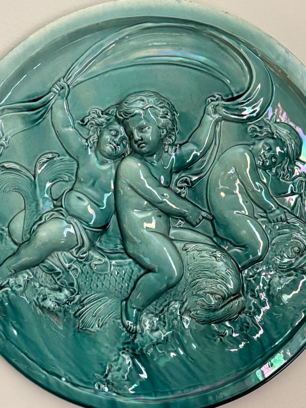 A circular turquoise-glazed ceramic plaque depicting cherubs sculpted in high relief holding flowing drapery. View V