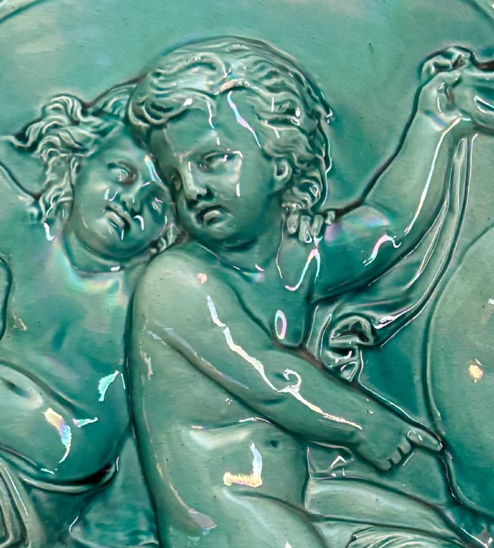 A circular turquoise-glazed ceramic plaque depicting cherubs sculpted in high relief holding flowing drapery. View VI
