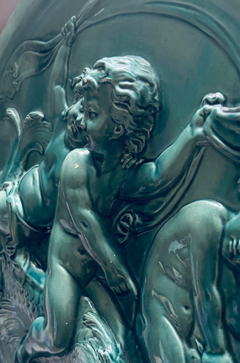 A circular turquoise-glazed ceramic plaque depicting cherubs sculpted in high relief holding flowing drapery. View VII
