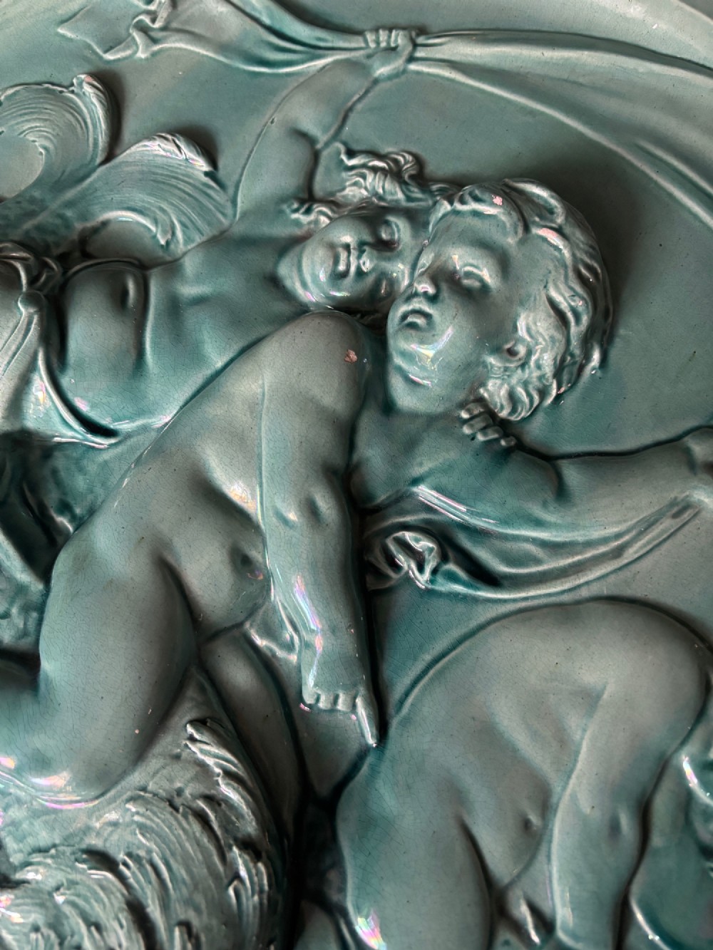 A circular turquoise-glazed ceramic plaque depicting cherubs sculpted in high relief holding flowing drapery. View X