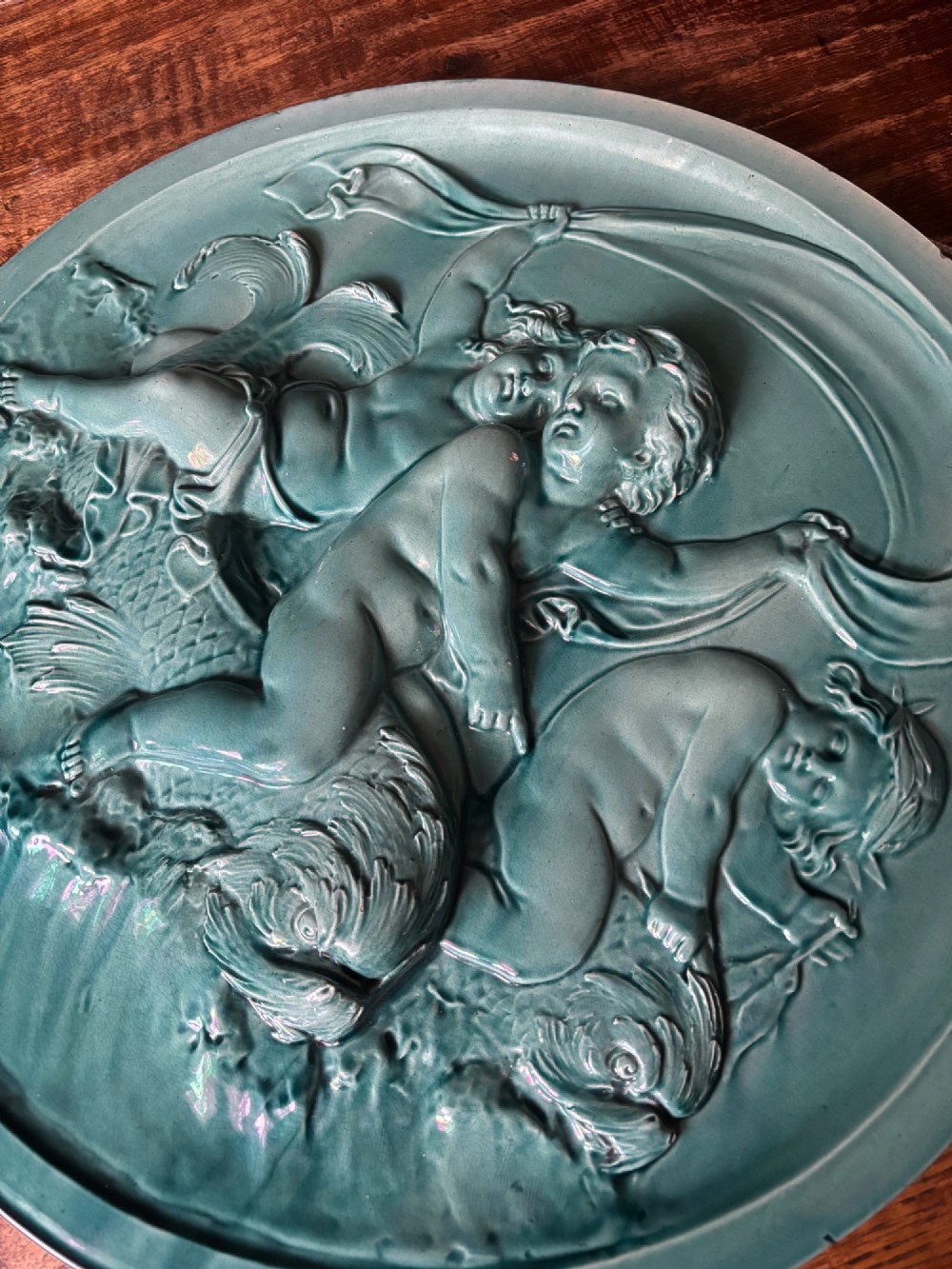 A circular turquoise-glazed ceramic plaque depicting cherubs sculpted in high relief holding flowing drapery. View XI