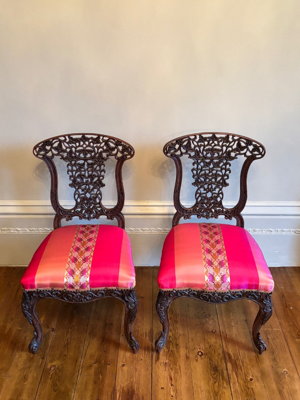 A pair of 19th-century Anglo-Indian padauk wood side chairs featuring intricately carved openwork backs, cabriole legs, and bright pink silk seat cushions. View II
