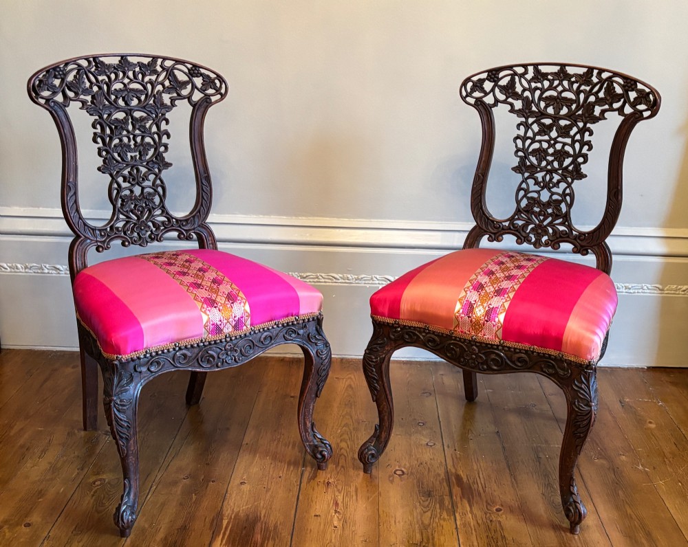 A pair of 19th-century Anglo-Indian padauk wood side chairs featuring intricately carved openwork backs, cabriole legs, and bright pink silk seat cushions. View V