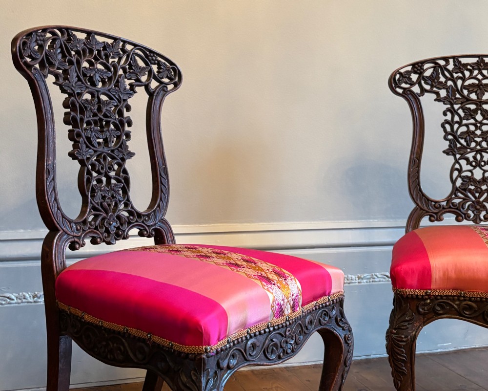 A pair of 19th-century Anglo-Indian padauk wood side chairs featuring intricately carved openwork backs, cabriole legs, and bright pink silk seat cushions. View VI