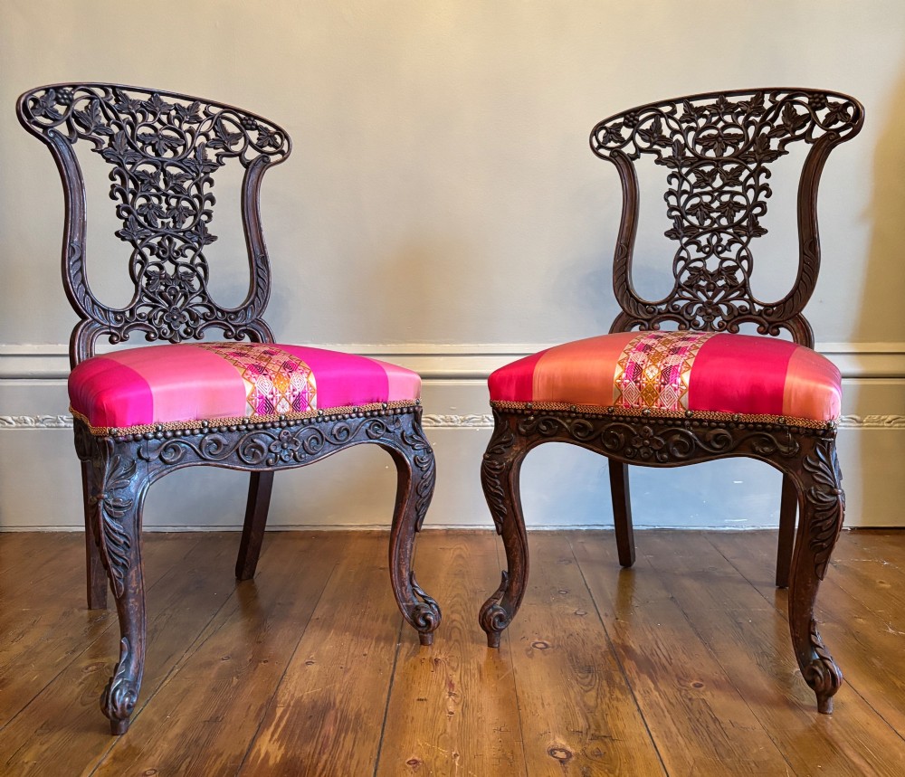 A pair of 19th-century Anglo-Indian padauk wood side chairs featuring intricately carved openwork backs, cabriole legs, and bright pink silk seat cushions. View VII