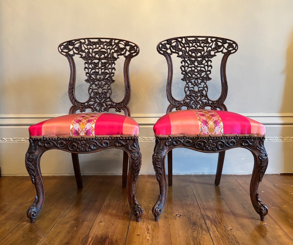 A pair of 19th-century Anglo-Indian padauk wood side chairs featuring intricately carved openwork backs, cabriole legs, and bright pink silk seat cushions.