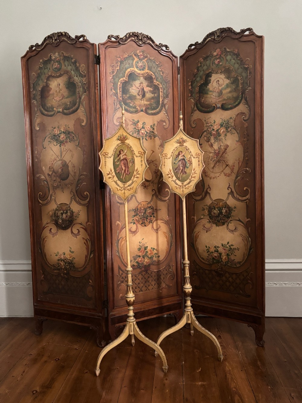 A pair of antique Georgian cream painted Hepplewhite pole screens featuring central oval vignettes depicting classical female figures with floral surround details on tripod spade-foot bases. View II
