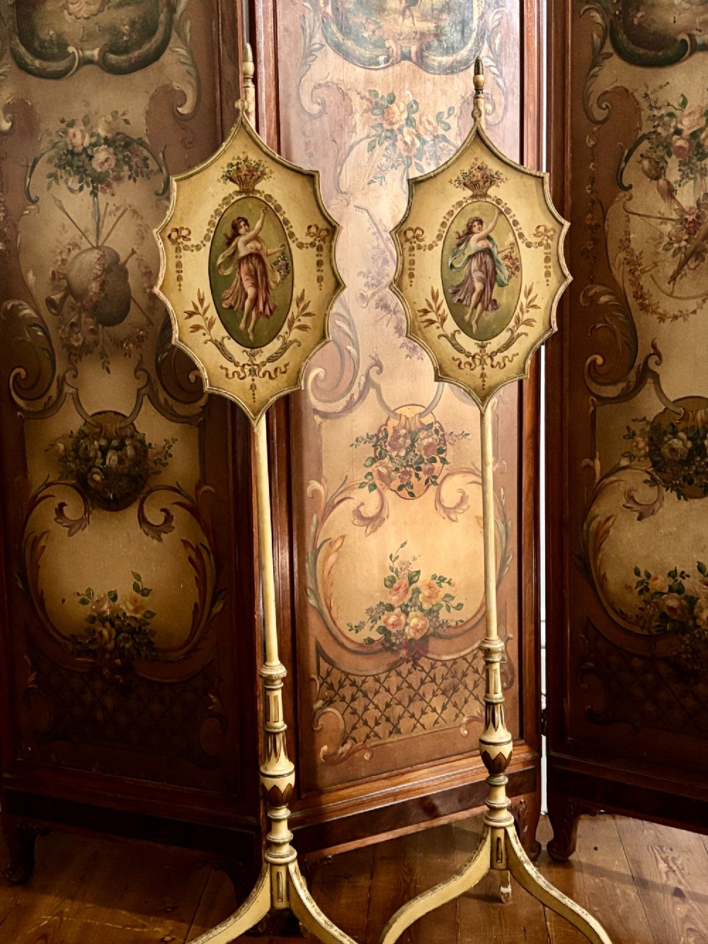 A pair of antique Georgian cream painted Hepplewhite pole screens featuring central oval vignettes depicting classical female figures with floral surround details on tripod spade-foot bases.