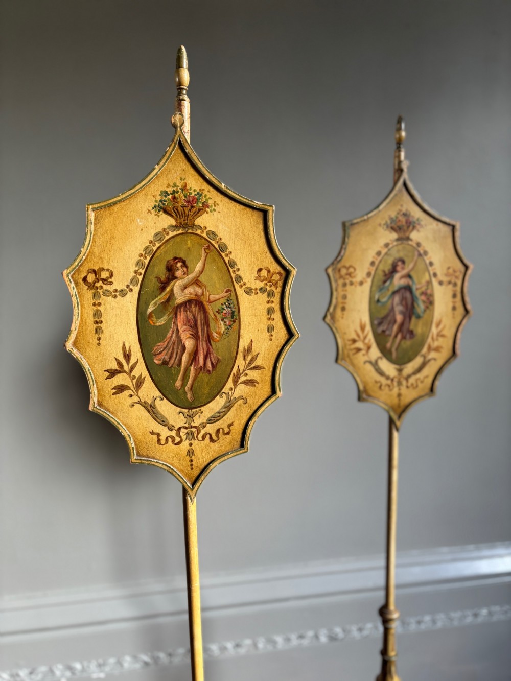 A pair of antique Georgian cream painted Hepplewhite pole screens featuring central oval vignettes depicting classical female figures with floral surround details on tripod spade-foot bases. View VII