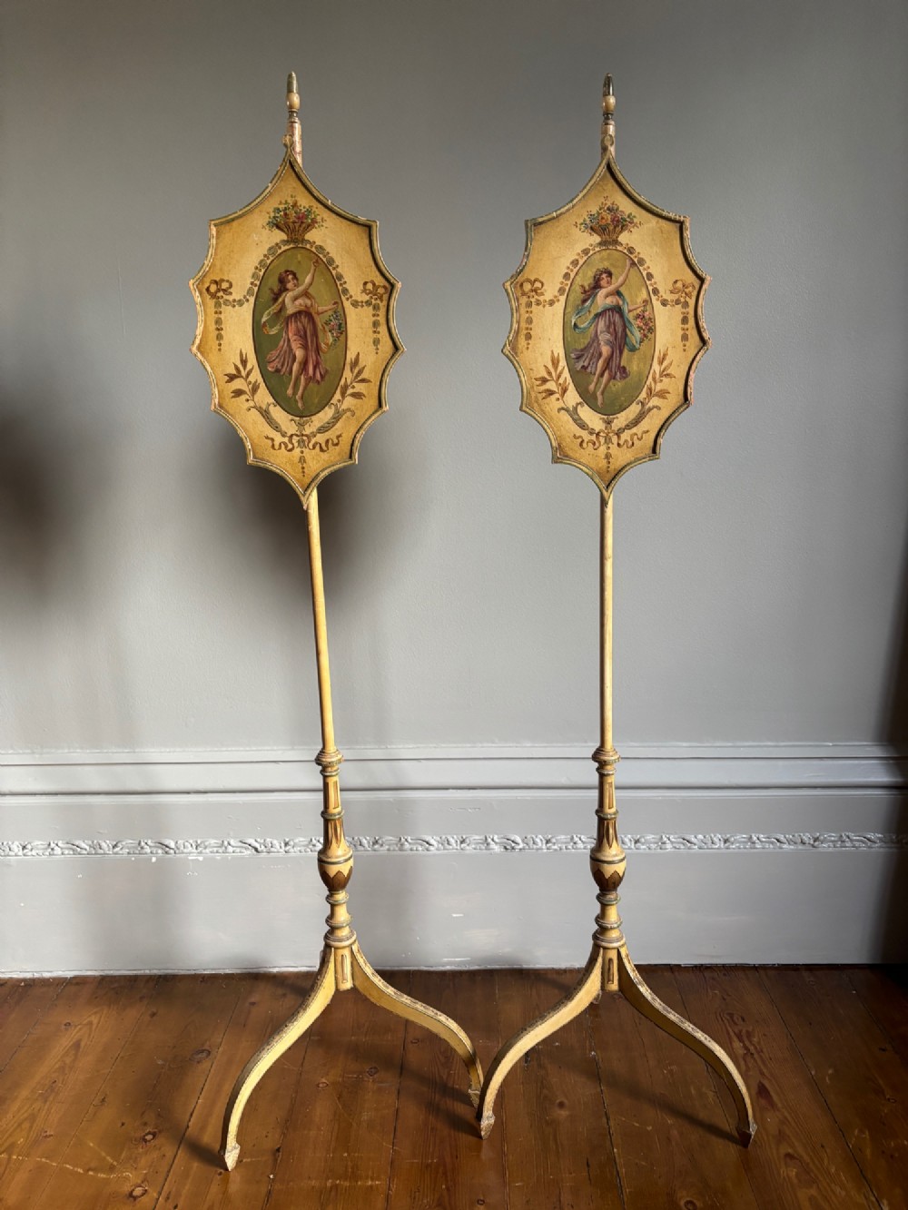 A pair of antique Georgian cream painted Hepplewhite pole screens featuring central oval vignettes depicting classical female figures with floral surround details on tripod spade-foot bases. View IX