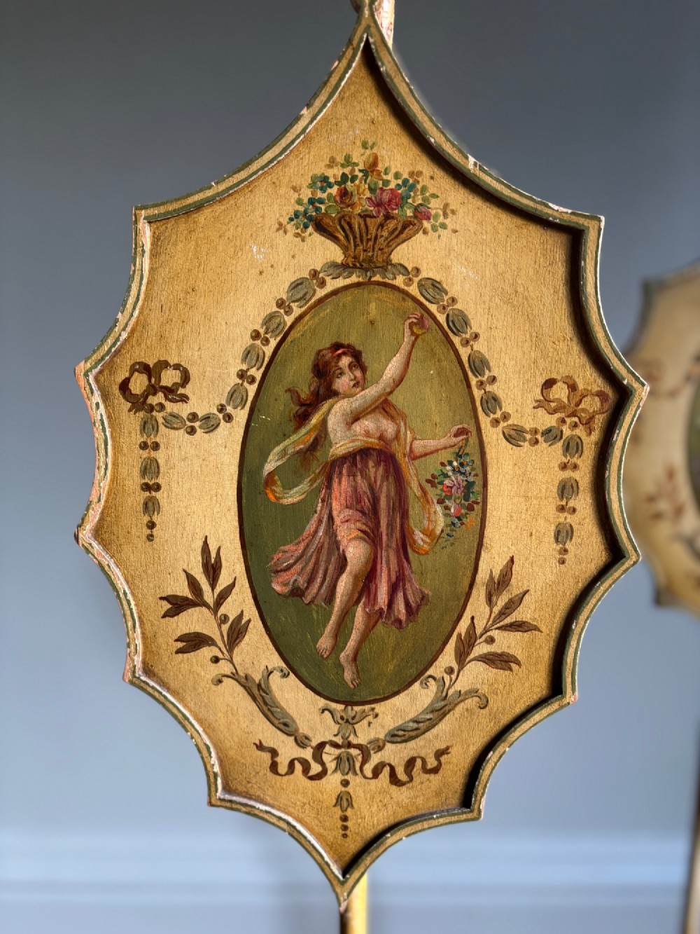 A pair of antique Georgian cream painted Hepplewhite pole screens featuring central oval vignettes depicting classical female figures with floral surround details on tripod spade-foot bases. View X