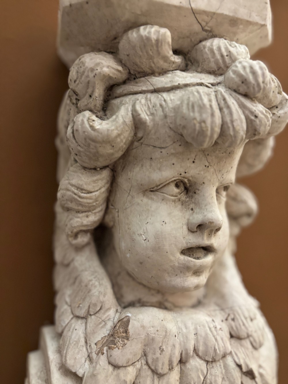 A carved off-white 19th-century wall corbel featuring a detailed winged putto head above layered acanthus leaves. View V