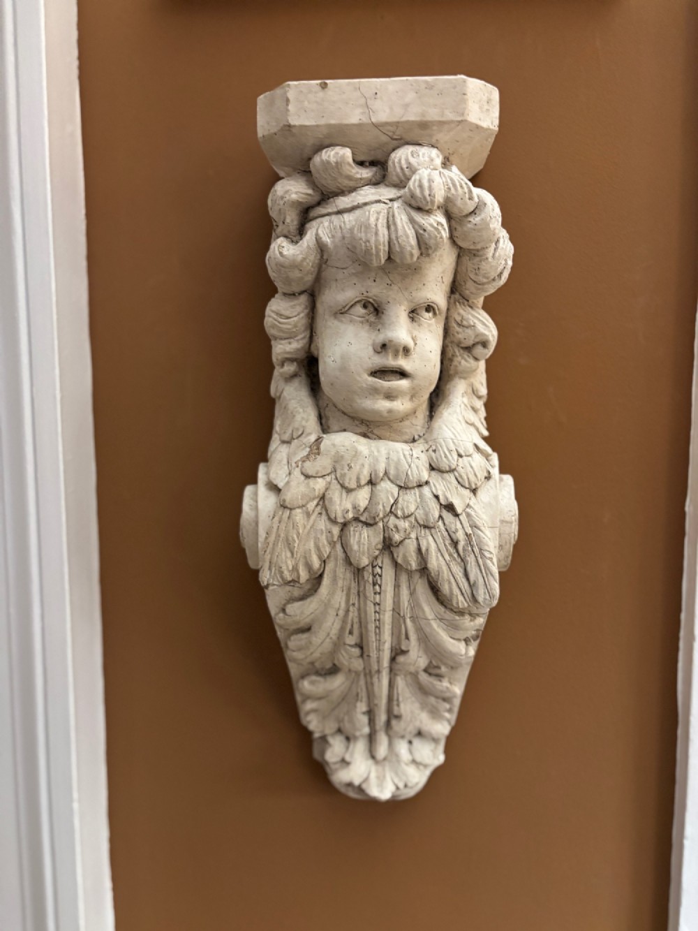 A carved off-white 19th-century wall corbel featuring a detailed winged putto head above layered acanthus leaves. View VII