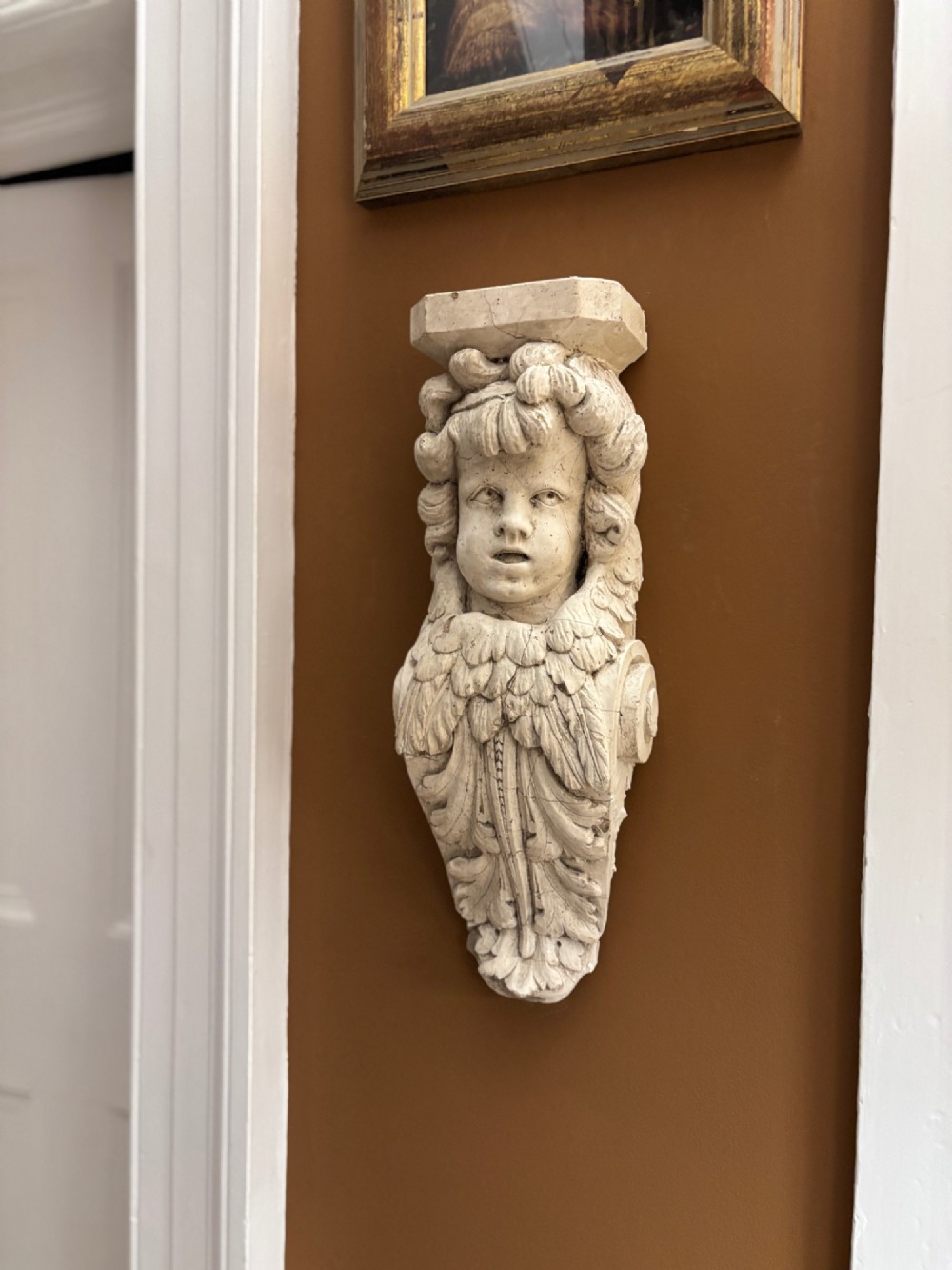 A carved off-white 19th-century wall corbel featuring a detailed winged putto head above layered acanthus leaves. View IX