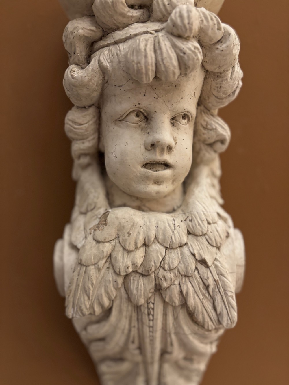 A carved off-white 19th-century wall corbel featuring a detailed winged putto head above layered acanthus leaves. View X
