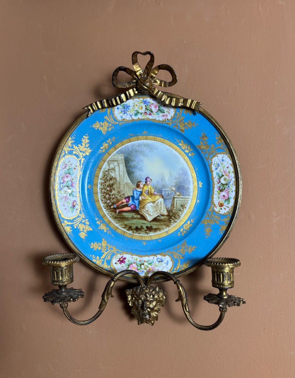 A circular sky blue porcelain plaque with a central hand-painted romantic couple and floral panels, framed in gilt bronze with a ribbon bow crest and two candle arms.