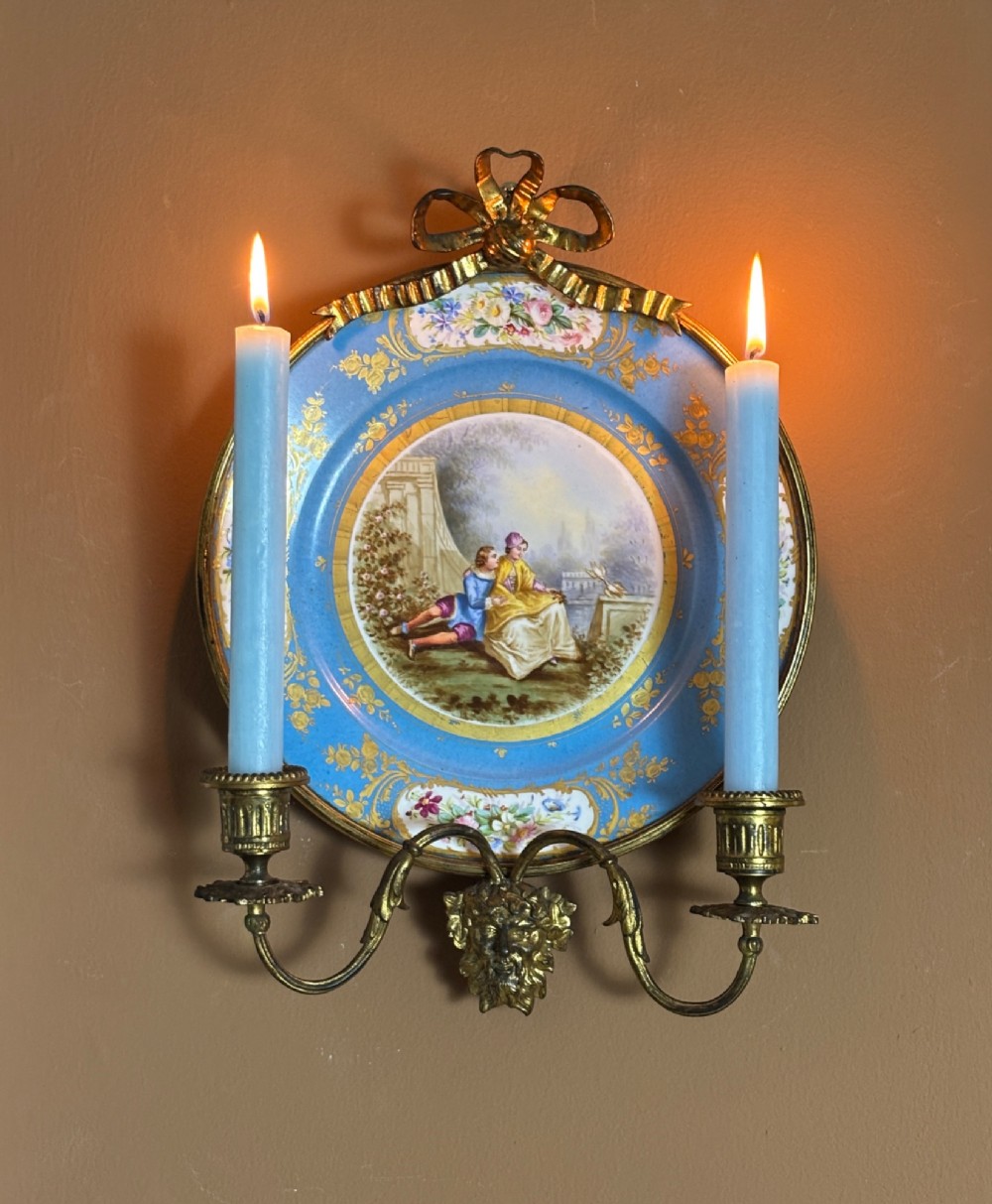 A circular sky blue porcelain plaque with a central hand-painted romantic couple and floral panels, framed in gilt bronze with a ribbon bow crest and two candle arms. View II