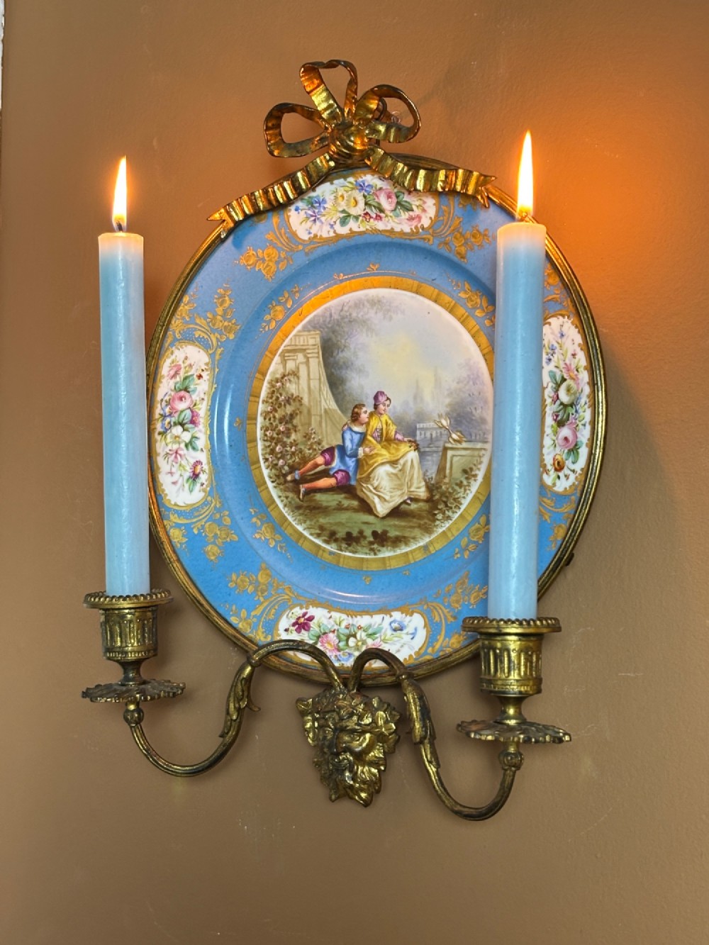 A circular sky blue porcelain plaque with a central hand-painted romantic couple and floral panels, framed in gilt bronze with a ribbon bow crest and two candle arms. View III