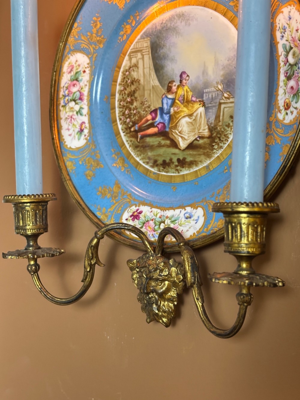 A circular sky blue porcelain plaque with a central hand-painted romantic couple and floral panels, framed in gilt bronze with a ribbon bow crest and two candle arms. View IV