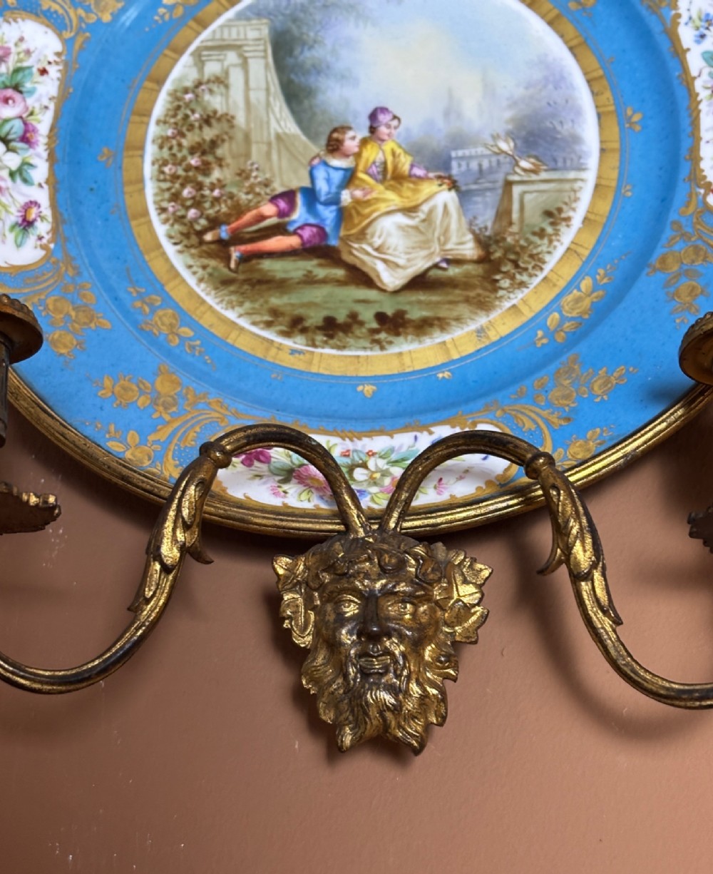 A circular sky blue porcelain plaque with a central hand-painted romantic couple and floral panels, framed in gilt bronze with a ribbon bow crest and two candle arms. View V