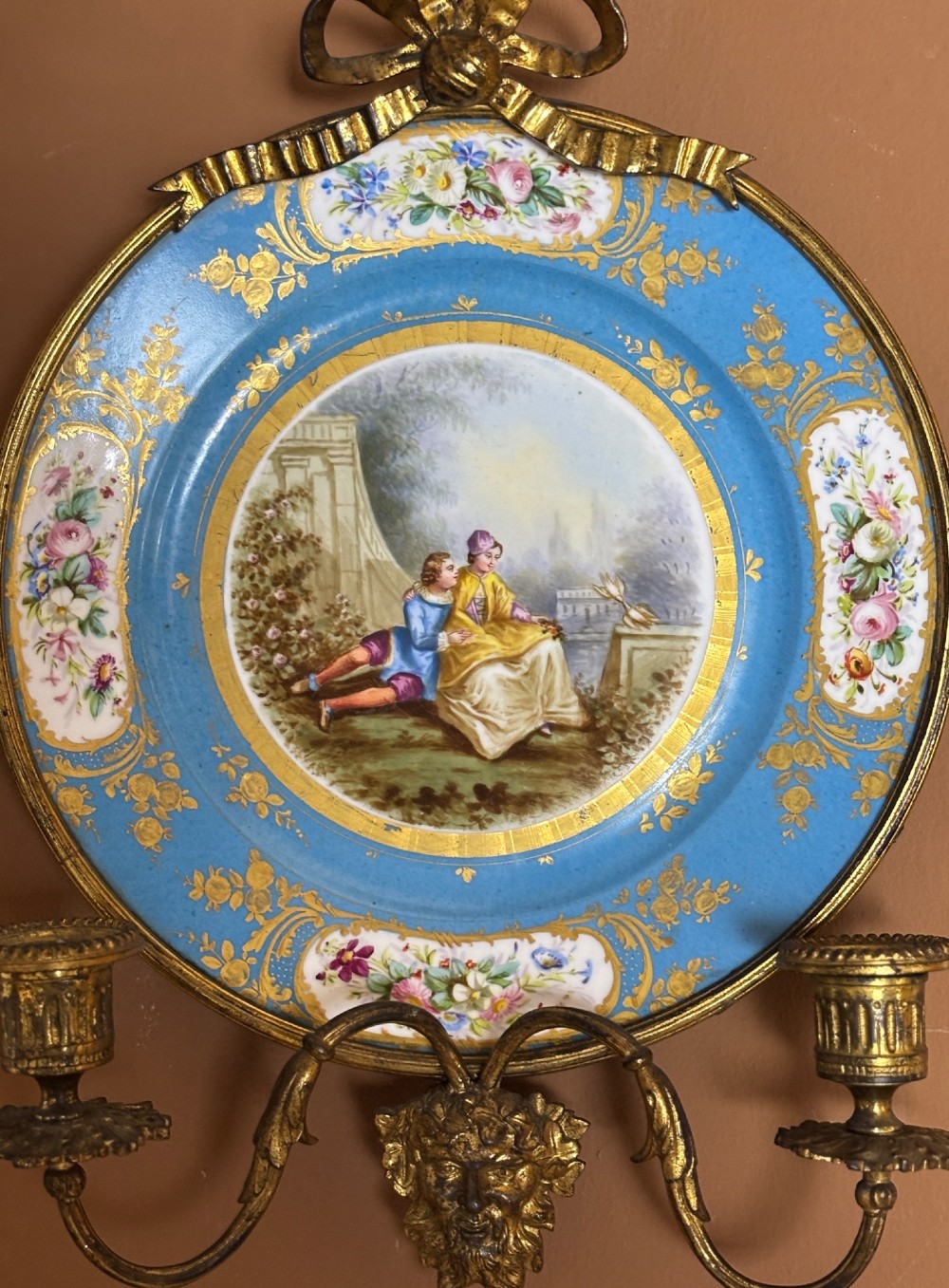 A circular sky blue porcelain plaque with a central hand-painted romantic couple and floral panels, framed in gilt bronze with a ribbon bow crest and two candle arms. View VI