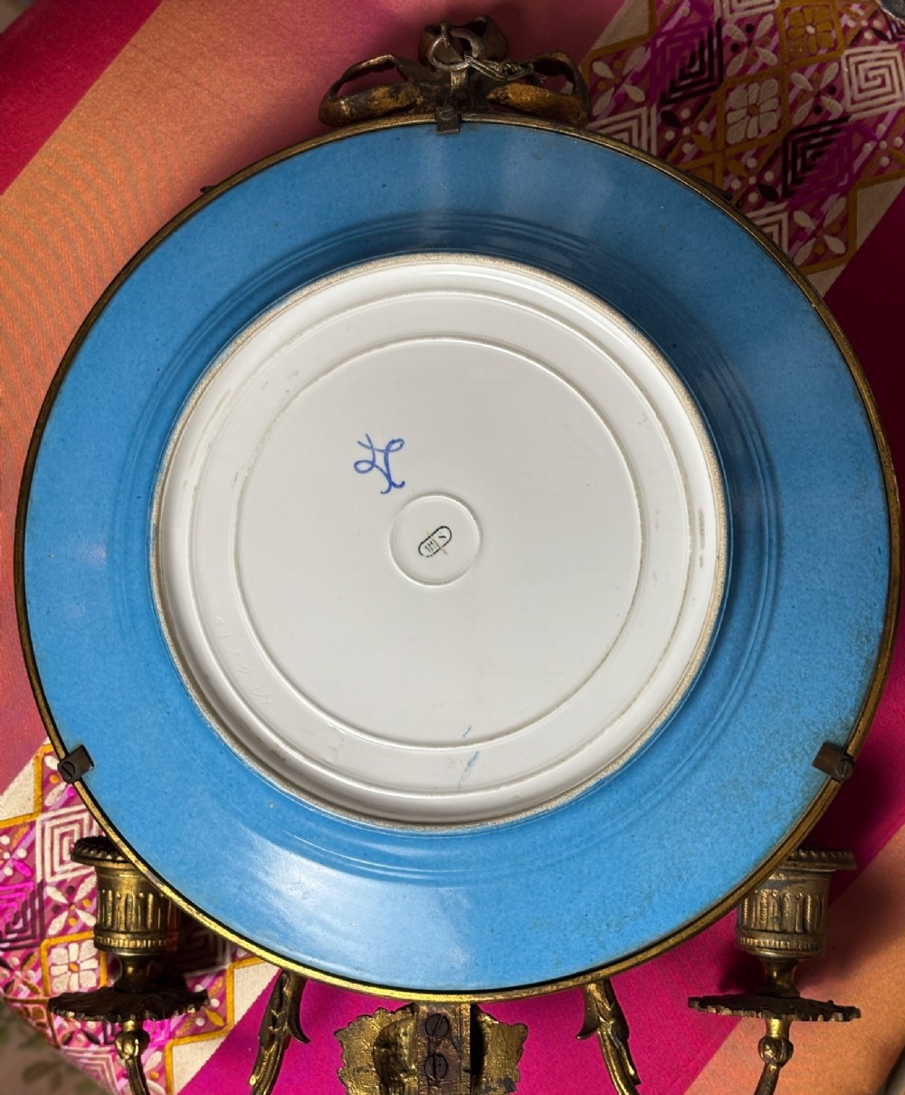 A circular sky blue porcelain plaque with a central hand-painted romantic couple and floral panels, framed in gilt bronze with a ribbon bow crest and two candle arms. View VII